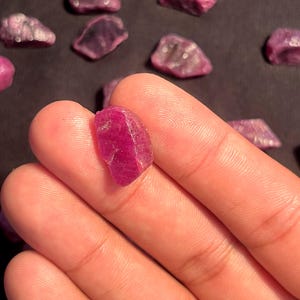Peut inclure: Une petite pierre précieuse rubis brute, d'un rouge rosé profond, avec une forme irrégulière et facettée. La pierre présente une qualité translucide avec des inclusions naturelles et une texture rugueuse non polie, sur un fond sombre.