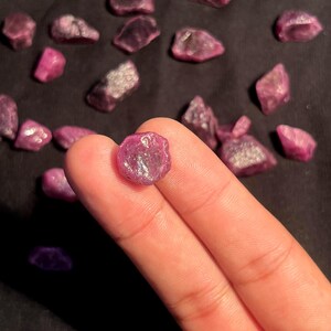 K&ouml;nnte beinhalten: Ein kleiner, roher, tief pink-roter Edelstein, der zwischen zwei Fingern gehalten wird, mit zahlreichen &auml;hnlichen ungeschliffenen, unregelm&auml;&szlig;igen, facettiert wirkenden Steinen im Hintergrund. Der mittlere Stein zeigt eine durchscheinende, kristalline Textur.