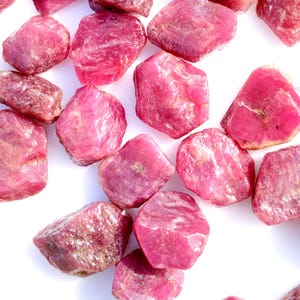May include: A selection of raw ruby gemstones in various sizes and shapes. The stones exhibit a spectrum of pink and red shades, with a natural, unpolished texture. The gemstones are scattered on a white surface.