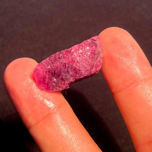 May include: A raw, unpolished ruby gemstone with a deep magenta-red hue and a textured, crystalline surface. The stone features natural facets and irregular edges, displaying a translucent quality that catches light across its uneven, jagged structure.