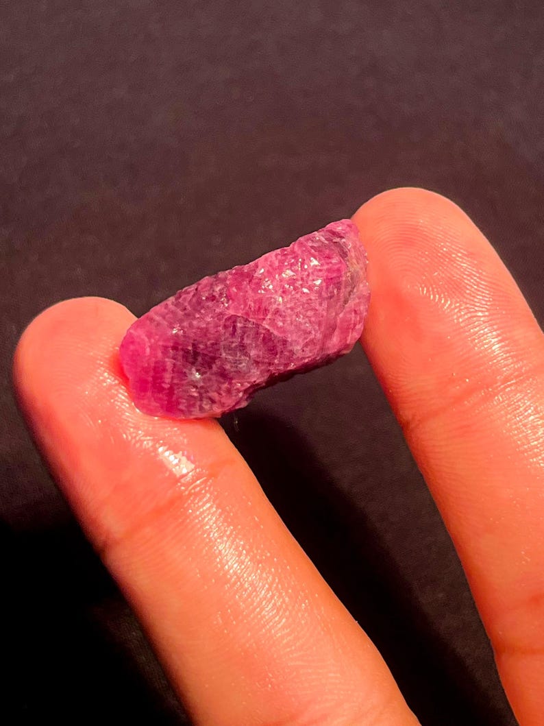 May include: A raw, unpolished ruby gemstone with a deep magenta-red hue and a textured, crystalline surface. The stone features natural facets and irregular edges, displaying a translucent quality that catches light across its uneven, jagged structure.