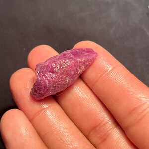 May include: A raw, unpolished ruby gemstone with a deep magenta-red hue. The stone features a rough, crystalline texture with natural facets and irregular edges, displaying a translucent quality that catches the light across its uneven surface.