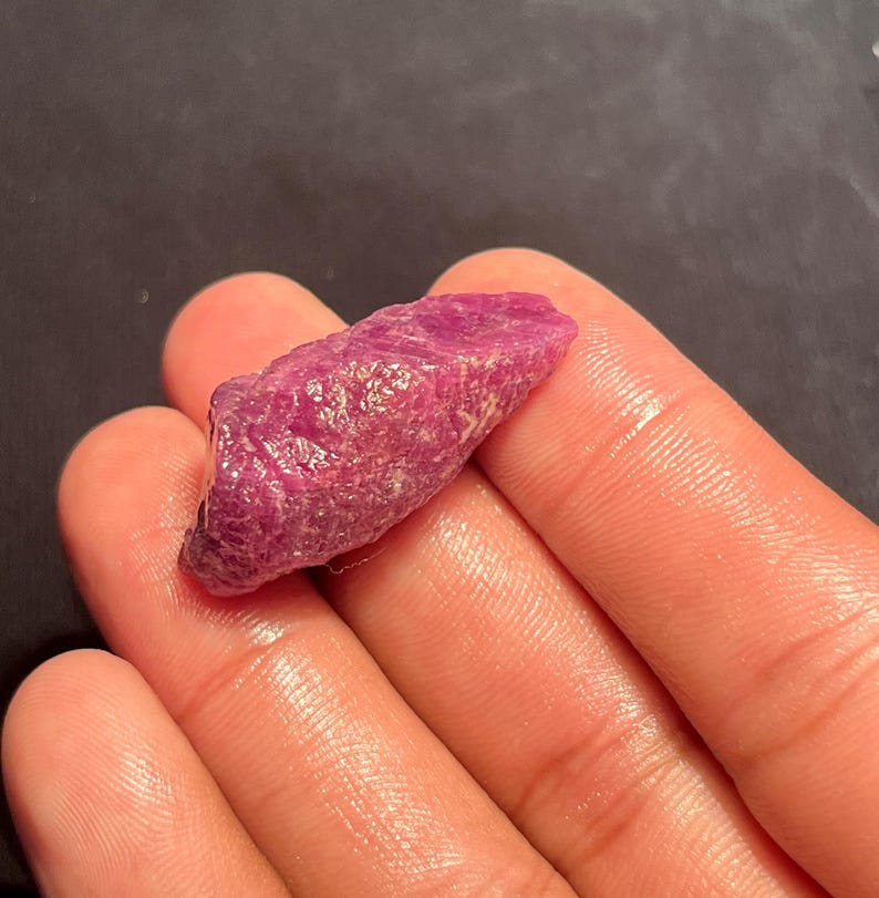 May include: A raw, unpolished ruby gemstone with a deep magenta-red hue. The stone features a rough, crystalline texture with natural facets and irregular edges, displaying a translucent quality that catches the light across its uneven surface.