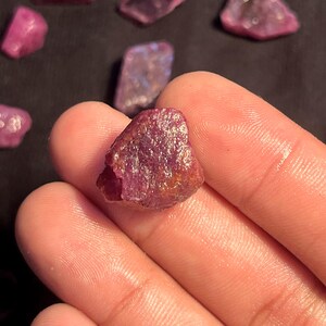 K&ouml;nnte beinhalten: Ein roher, unpolierter Rubin-Edelstein mit einem tiefen Magenta- und r&ouml;tlich-violetten Farbton. Der Stein weist eine raue, texturierte Oberfl&auml;che mit nat&uuml;rlichen kristallinen Facetten und unregelm&auml;&szlig;igen Kanten auf, die Farbvariationen zeigen.