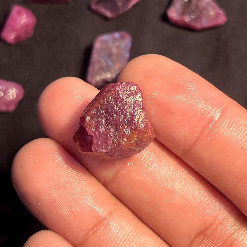 K&ouml;nnte beinhalten: Ein roher, unpolierter Rubin-Edelstein mit einem tiefen Magenta- und r&ouml;tlich-violetten Farbton. Der Stein weist eine raue, texturierte Oberfl&auml;che mit nat&uuml;rlichen kristallinen Facetten und unregelm&auml;&szlig;igen Kanten auf, die Farbvariationen zeigen.