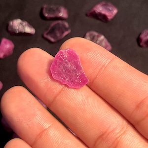 May include: A small, raw, vibrant pinkish-red gemstone fragment with an irregular, jagged shape and a translucent, crystalline texture. The stone displays natural facets and internal inclusions, held against a dark, out-of-focus background.