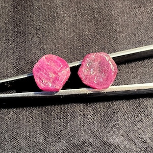 Puede incluir: Dos rubíes en bruto de color rojo rosado vibrante con superficies irregulares y facetadas, sostenidos por unas pinzas de metal plateado. Las piedras preciosas muestran una calidad translúcida con inclusiones visibles y textura natural.