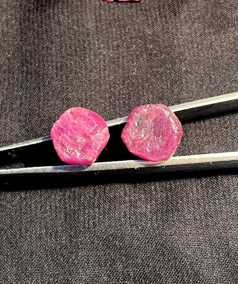 K&ouml;nnte beinhalten: Zwei rohe, leuchtend pink-rote Rubine mit unregelm&auml;&szlig;igen, facettierten Oberfl&auml;chen, gehalten von einer silberfarbenen Metallpinzette. Die Edelsteine weisen eine durchscheinende Qualit&auml;t mit sichtbaren Einschl&uuml;ssen und eine nat&uuml;rliche Textur auf.