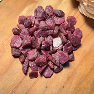 May include: A collection of raw, unpolished ruby gemstones in various shades of deep pink and red. These small, irregular mineral fragments feature natural textures and opaque surfaces, clustered together in a dense, organic pile.