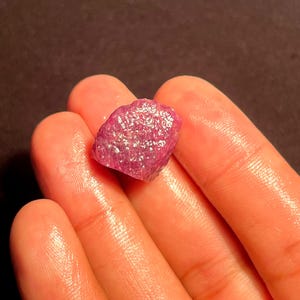May include: A raw, unpolished gemstone with a vibrant magenta-pink hue and a textured, crystalline surface. The stone features an irregular, multifaceted shape with natural indentations and a semi-translucent quality that catches the light.