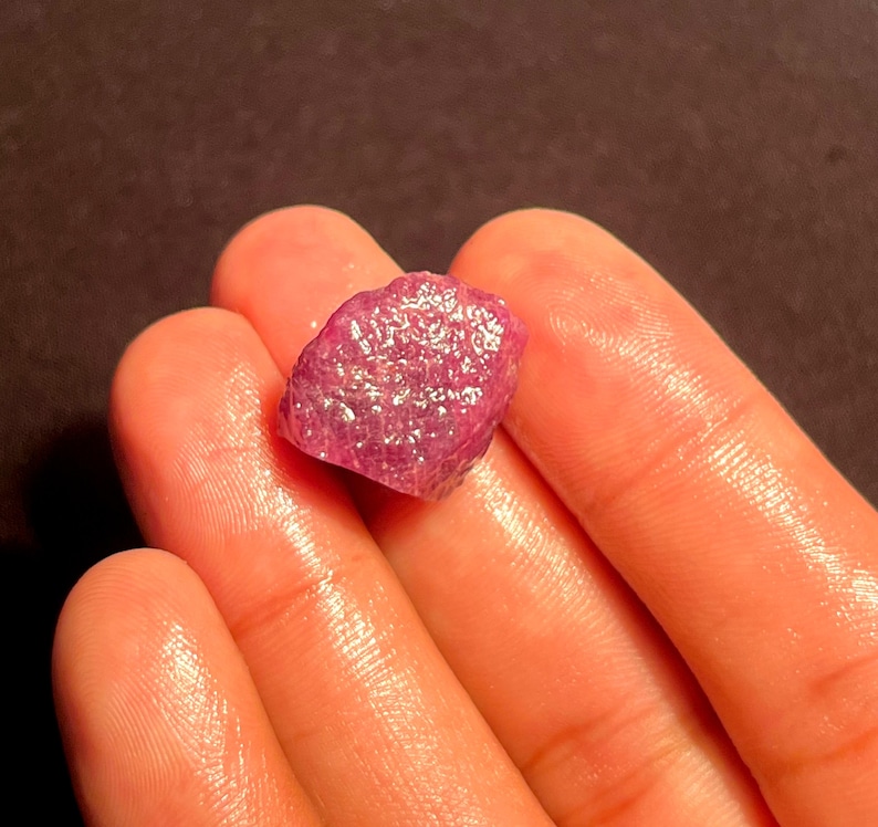 May include: A raw, unpolished gemstone with a vibrant magenta-pink hue and a textured, crystalline surface. The stone features an irregular, multifaceted shape with natural indentations and a semi-translucent quality that catches the light.