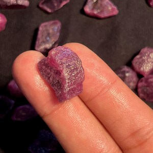 May include: A raw, unpolished ruby gemstone with a deep magenta and purple hue. The stone features a rough, crystalline texture with natural facets and internal inclusions, displaying a translucent quality when light hits the irregular surface.