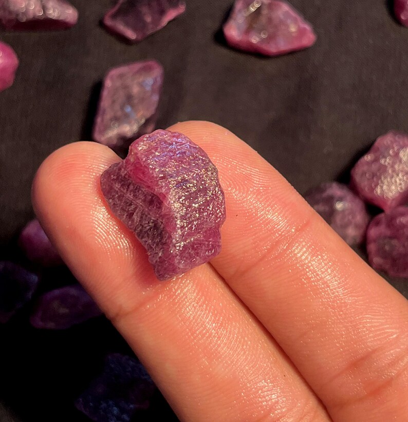 May include: A raw, unpolished ruby gemstone with a deep magenta and purple hue. The stone features a rough, crystalline texture with natural facets and internal inclusions, displaying a translucent quality when light hits the irregular surface.