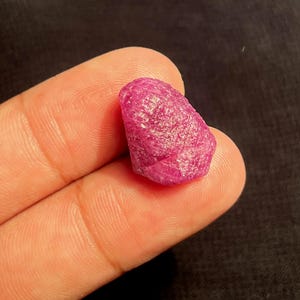 May include: A raw, unpolished gemstone with a vibrant, deep pinkish-red hue. The stone features a naturally textured, crystalline surface with irregular facets and edges, showcasing a translucent quality that catches light across its uneven form.