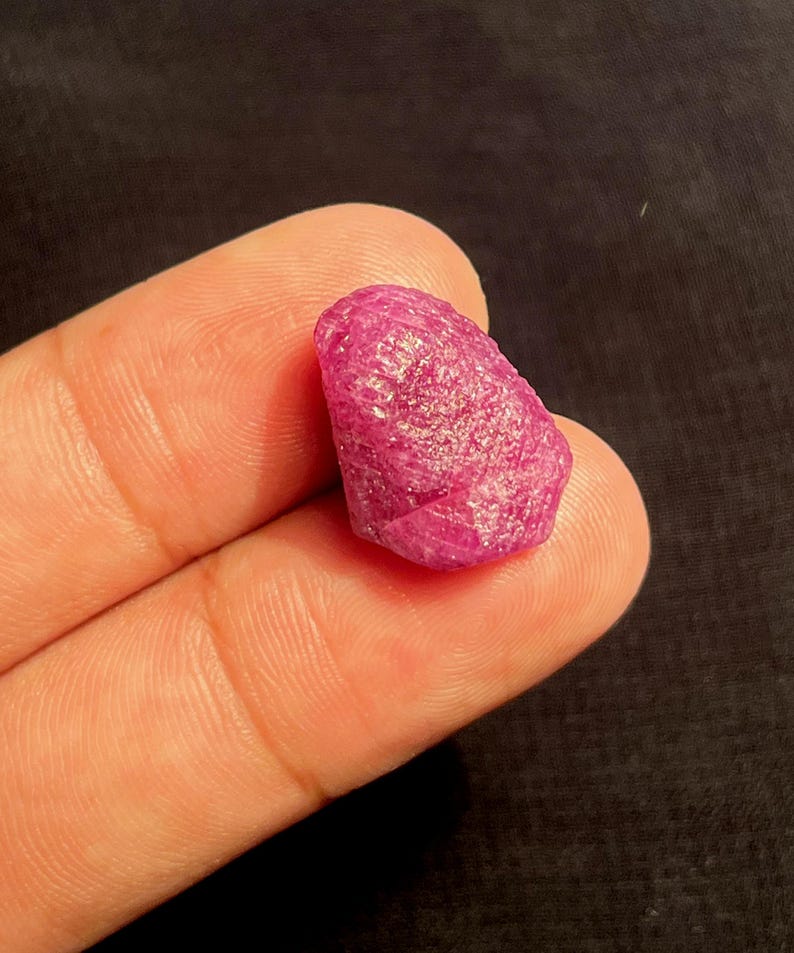 May include: A raw, unpolished gemstone with a vibrant, deep pinkish-red hue. The stone features a naturally textured, crystalline surface with irregular facets and edges, showcasing a translucent quality that catches light across its uneven form.