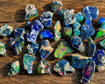 Könnte beinhalten: Eine Sammlung roher, unpolierter Opalsteine mit tiefblauen, grünen und irisierenden Farbblitzen. Jeder Stein weist eine raue, natürliche Textur mit unterschiedlichen Formen und Größen auf und zeigt lebhaftes Farbspiel vor dunklem, felsigem Matrixmaterial.