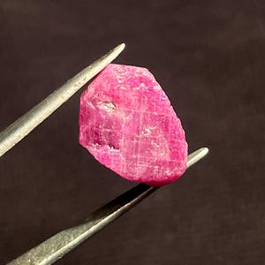 May include: A raw, unpolished ruby gemstone with a vibrant, saturated pinkish-red hue. The stone features a natural, irregular shape with visible crystalline surface textures, facets, and internal inclusions held by metal tweezers.