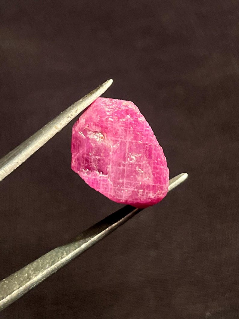 May include: A raw, unpolished ruby gemstone with a vibrant, saturated pinkish-red hue. The stone features a natural, irregular shape with visible crystalline surface textures, facets, and internal inclusions held by metal tweezers.
