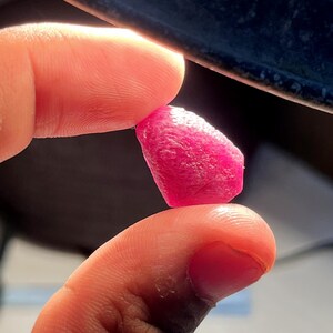 May include: A small, raw, vibrant pinkish-red gemstone with a textured, irregular surface. The stone is translucent, allowing light to pass through its crystalline structure, highlighting its deep, saturated color and natural, unpolished, faceted shape.