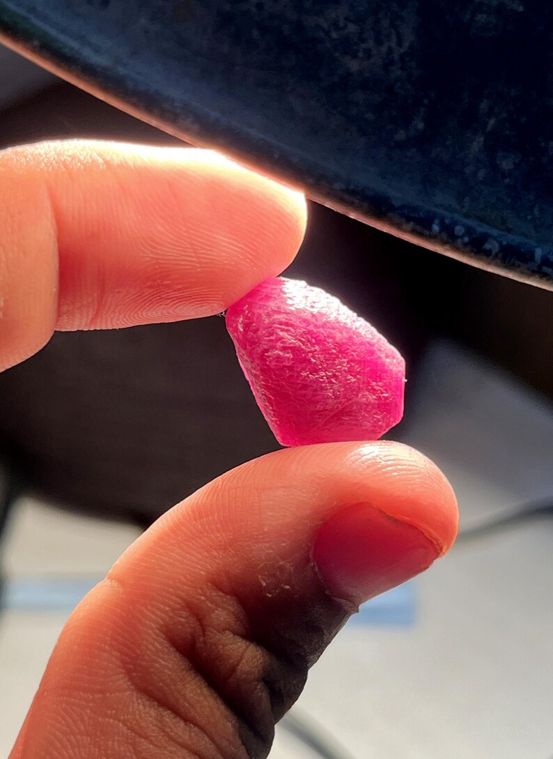 May include: A small, raw, vibrant pinkish-red gemstone with a textured, irregular surface. The stone is translucent, allowing light to pass through its crystalline structure, highlighting its deep, saturated color and natural, unpolished, faceted shape.