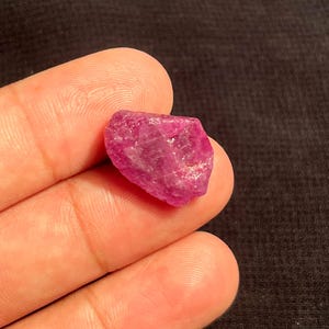 May include: A raw, unpolished gemstone with a vibrant magenta-pink hue. The stone features an irregular, multifaceted shape with a semi-translucent texture, displaying natural crystalline structures and internal light reflections across its surface.