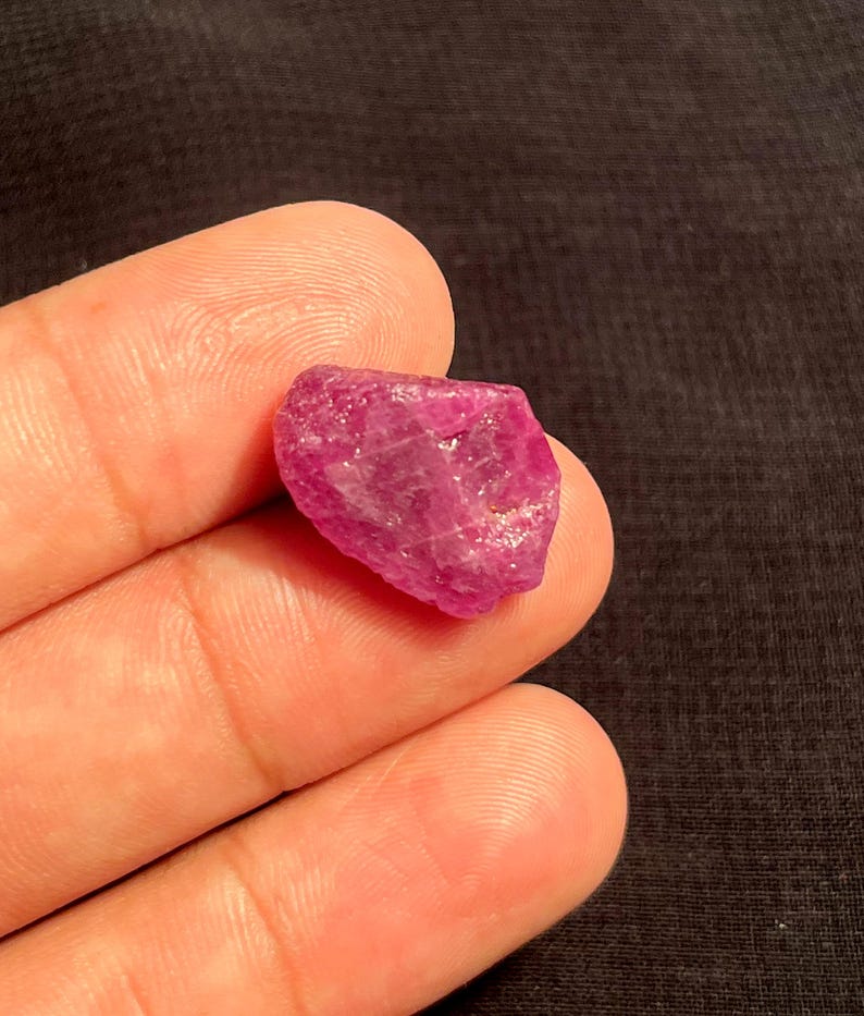 May include: A raw, unpolished gemstone with a vibrant magenta-pink hue. The stone features an irregular, multifaceted shape with a semi-translucent texture, displaying natural crystalline structures and internal light reflections across its surface.