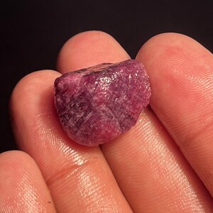 May include: A raw, unpolished ruby gemstone with a deep magenta and dark red color. The stone features a rough, textured surface with visible crystalline structures and natural facets, displaying a mix of opaque and translucent areas.