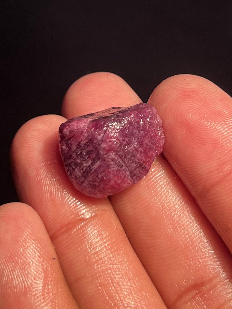 May include: A raw, unpolished ruby gemstone with a deep magenta and dark red color. The stone features a rough, textured surface with visible crystalline structures and natural facets, displaying a mix of opaque and translucent areas.