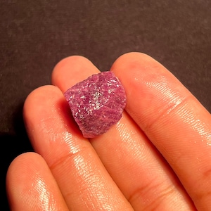 May include: A raw, unpolished gemstone with a deep magenta and reddish-purple hue. The stone features a rough, textured surface with natural crystalline facets and irregular edges, displaying a translucent quality when light hits the surface.
