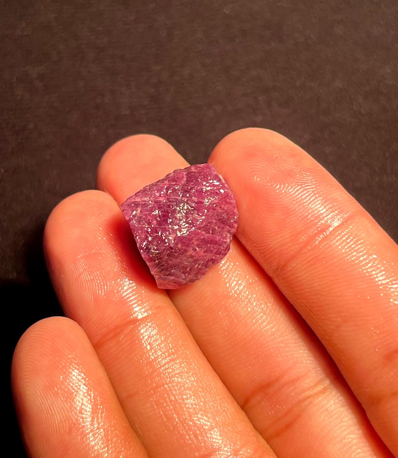 May include: A raw, unpolished gemstone with a deep magenta and reddish-purple hue. The stone features a rough, textured surface with natural crystalline facets and irregular edges, displaying a translucent quality when light hits the surface.