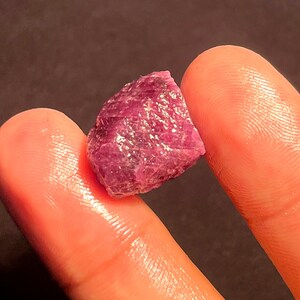 May include: A raw, unpolished ruby gemstone with a deep magenta and purple hue. The stone features a jagged, crystalline surface with natural facets and an irregular, angular shape that reflects light across its textured, translucent structure.