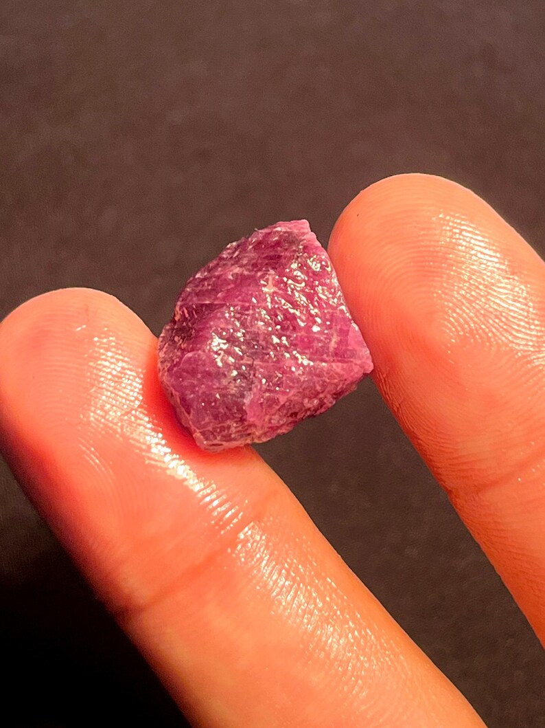 May include: A raw, unpolished ruby gemstone with a deep magenta and purple hue. The stone features a jagged, crystalline surface with natural facets and an irregular, angular shape that reflects light across its textured, translucent structure.