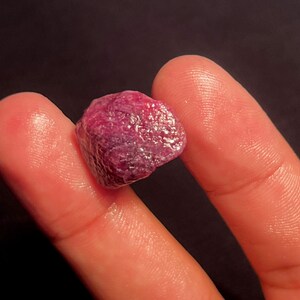 May include: A raw, unpolished ruby gemstone with a deep magenta-red color and a textured, multifaceted surface. The stone displays natural crystalline formations and opaque characteristics, showcasing a vibrant, saturated hue throughout its structure.