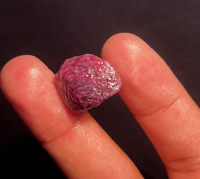 May include: A raw, unpolished ruby gemstone with a deep magenta-red color and a textured, multifaceted surface. The stone displays natural crystalline formations and opaque characteristics, showcasing a vibrant, saturated hue throughout its structure.