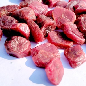 May include: A pile of raw ruby gemstones, showcasing a deep red colour with variations in tone. The stones are of different sizes and shapes, with a rough, natural texture. The rubies are displayed against a white backdrop.