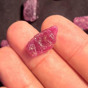 May include: A raw, unpolished ruby gemstone with a deep magenta-pink hue and a textured, irregular surface. The stone displays natural facets and crystalline structures, showcasing a vibrant, translucent quality throughout its jagged, organic form.