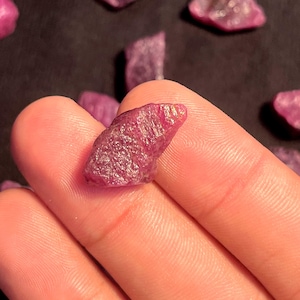 May include: A raw, unpolished ruby gemstone with a deep magenta-red hue and a textured, crystalline surface. The stone displays natural facets and irregular edges, showcasing a vibrant, opaque color profile typical of untreated mineral specimens.