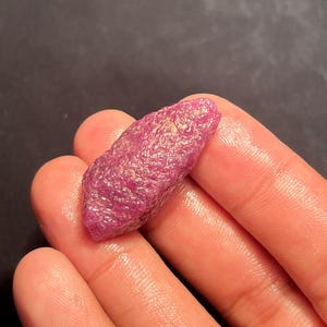 May include: A raw, unpolished ruby gemstone with a deep magenta-pink hue and a textured, crystalline surface. The stone has an elongated, irregular shape with natural facets and a slightly translucent appearance, showcasing its organic mineral structure.