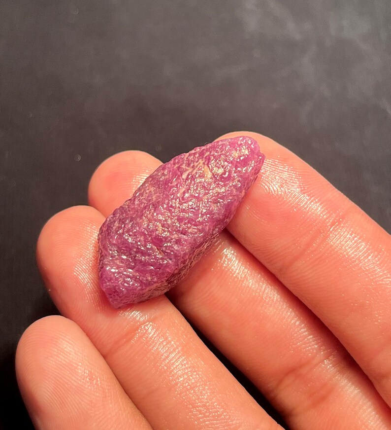 May include: A raw, unpolished ruby gemstone with a deep magenta-pink hue and a textured, crystalline surface. The stone has an elongated, irregular shape with natural facets and a slightly translucent appearance, showcasing its organic mineral structure.