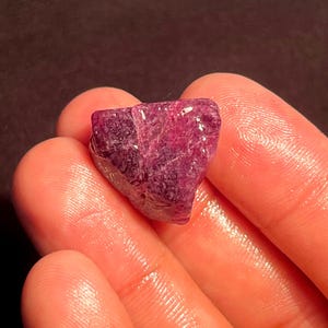 Peut inclure: Une pierre précieuse brute de couleur magenta profond avec une forme irrégulière à facettes. La pierre présente une qualité translucide avec des structures cristallines internes visibles et une texture de surface polie et réfléchissante.