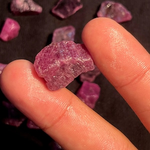 May include: A raw, unpolished ruby gemstone with a deep magenta-pink hue and a textured, crystalline surface. The stone is irregular in shape, featuring natural facets and inclusions, displayed alongside several other similar loose ruby pieces.