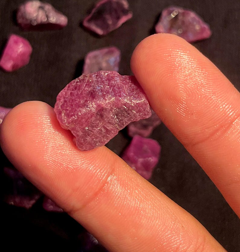May include: A raw, unpolished ruby gemstone with a deep magenta-pink hue and a textured, crystalline surface. The stone is irregular in shape, featuring natural facets and inclusions, displayed alongside several other similar loose ruby pieces.