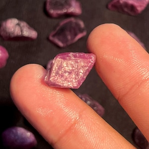 Peut inclure: Une petite pierre précieuse brute de couleur magenta, de forme irrégulière et facettée, présentant des structures cristallines internes visibles. La pierre est translucide avec une surface polie, sur un fond sombre avec d'autres pierres.