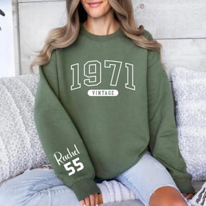 May include: Olive green sweatshirt with the year "1971" in white block letters and the word "VINTAGE" underneath. The sleeve features the name "Rachel" and the number "55" in white. The sweatshirt is a casual style.