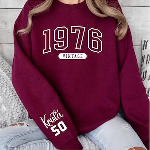 May include: A maroon sweatshirt with the year "1976" in white block letters and the word "VINTAGE" underneath. The sleeve has the name "Krista" and the number "50" in white script.