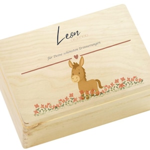 Puede incluir: Una caja de madera de color claro con tapa con bisagras. La tapa presenta un burro de dibujos animados, flores rojas y el nombre "Leon" en escritura cursiva. El texto en alemán dice "für Deine schönsten Erinnerungen."