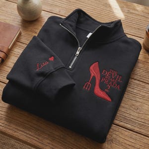 May include: Black quarter-zip sweatshirt with red embroidered design. Features the text "The Devil Wears Prada 2" and a high-heeled shoe with a trident. The left sleeve has the name "Lisa" and a heart.