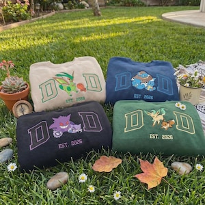 Puede incluir: Cuatro sudaderas de cuello redondo en beige, azul marino, negro y verde, cada una con un diseño de Pokémon bordado diferente. Las sudaderas presentan las letras "D D" y el texto "EST. 2026" o "EST. 2028". Las sudaderas están dispuestas sobre una superficie cubierta de hierba.