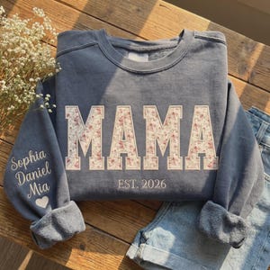 Könnte beinhalten: Ein blau-graues Sweatshirt mit dem Wort "MAMA" in großen Buchstaben mit floralem Muster auf der Vorderseite. Der Text "EST. 2026" steht darunter. Auf dem linken Ärmel sind die Namen "Sophia Daniel Mia" in Weiß gestickt, mit einem weißen Herz.