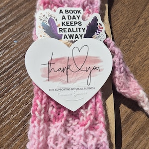 May include: A pink and white variegated knit bookmark attached to a cardboard backing. It features a heart-shaped sticker that says "thank you" and "FOR SUPPORTING MY SMALL BUSINESS" and a sticker that says "A BOOK A DAY KEEPS REALITY AWAY".